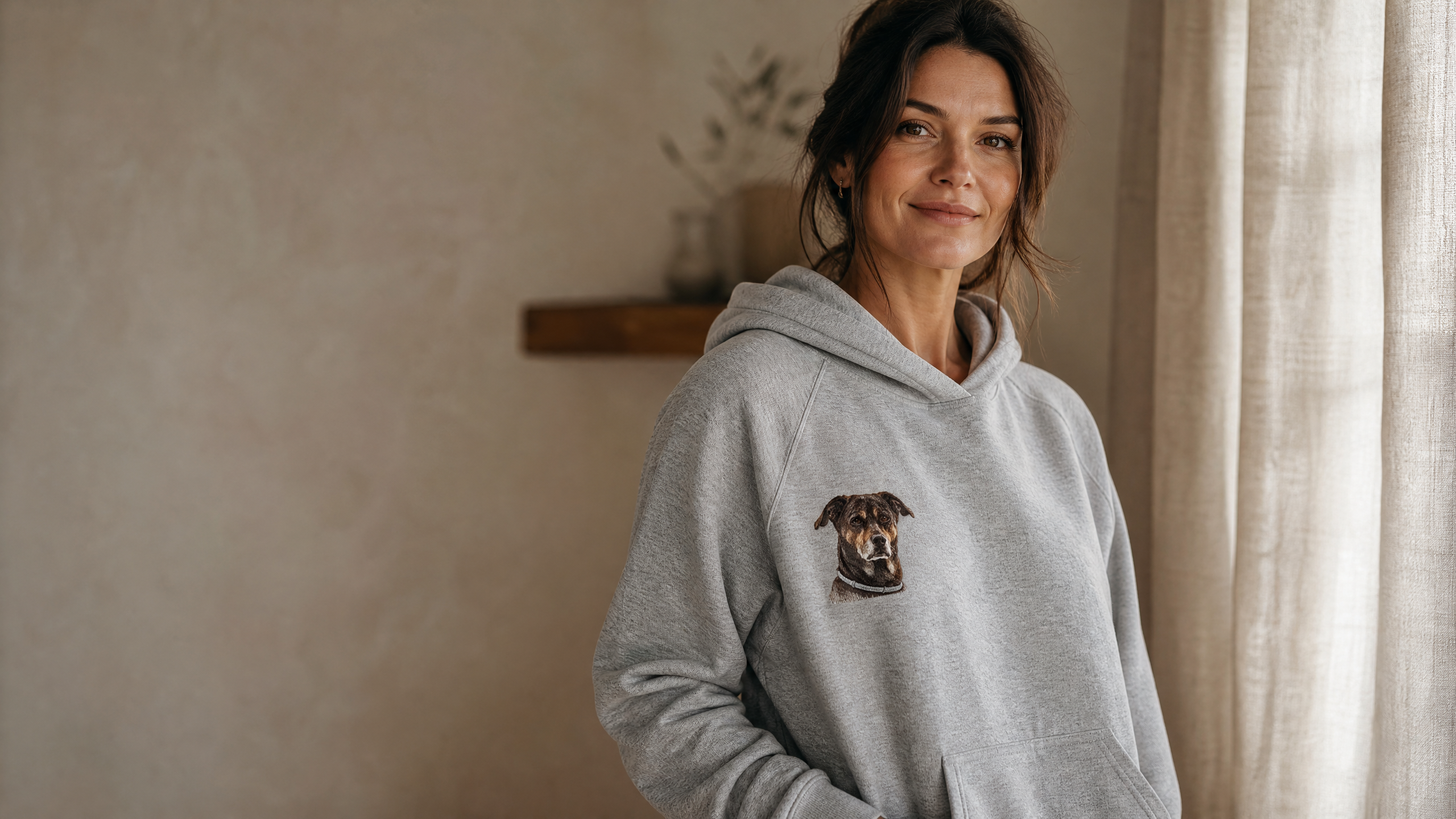 A woman wearing a heather-grey hoodie with a small embroidered portrait of a dark brown dog on the left chest